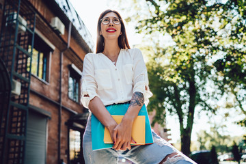 Millennial cheerful female student with sketch- and textbook for education in hands smiling at urban setting, happy woman in classic eyewear for vision protection standing at street and feeling good