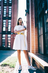 Full length portrait of beautiful teenage female model looking at camera during photo session standing at urbanity near buildings, trendy dressed Latino hipster girl posing during sunny summer day