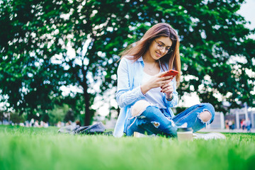 Young woman blogger in spectacles checking mail and sending messages via smartphone sitting in park, millennial woman student reading news from social networks using mobile phone and 4G in campus