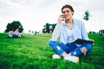 Attractive caucasian woman student sitting with coffee to go in park pondering on homework task, pensive female hipster in spectacles thinking about creative article recreating on green grass outdoors © BullRun