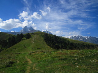 Fototapeta premium Mountain landscape of Svaneti on bright summer sunny day. Mountain lake, hills covered green grass on snowy rocky mountains background. Caucasus peaks in Georgia. Amazing view on wild georgian nature