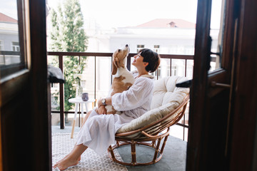 Relaxed young woman in white cozy bathrobe sitting with beagle dog on balcony in summer morning. Pretty brunette girl in wristwatch holding puppy on knees and smiling.