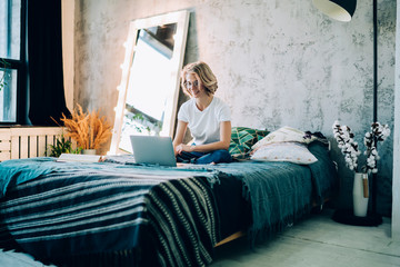 Caucasian female freelancer sitting at comfortable bed in cosy home interior and working remotely on modern laptop computer, happy woman in spectacles enjoying online shopping in web store