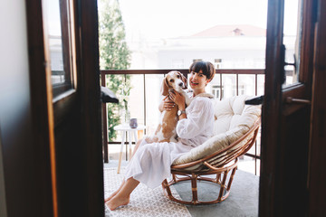 Barefooted laughing girl looking at camera, holding beagle dog while sitting in cozy chair. Lovely...