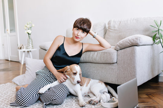 Graceful brown-haired girl in black tank-top relaxing on carpet near striped cushions and stroking beagle puppy. Indoor portrait of joyful young woman posing with pleasure while playing with dog.