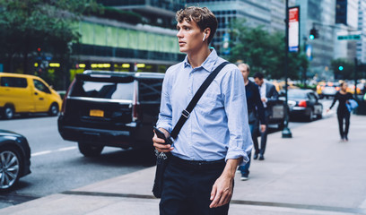 Pensive male manager with briefcase walking to corporate enterprise and listening audio book via electronic headphones,employer in earbuds thinking on received information in voice message on cellular