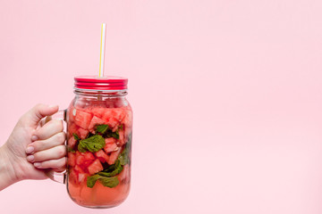 Young woman hands holding freshly squeezed watermelon lemonade of citrus fruits. Female with mason jar full of cold cocktail, mint leaves. Pink background, copy space, close up