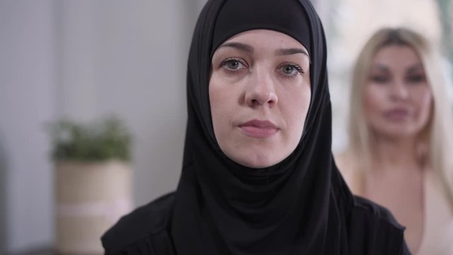Change of focus from Caucasian blond woman to face of Muslim lady in hijab looking at camera. Modern girl and conservative eastern woman posing indoors. Traditions, culture, diversity.