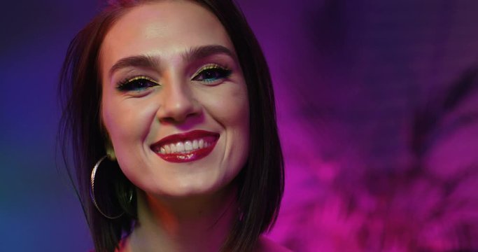 Close up of beautiful Caucasian young woman with short dark hair and bright make up smiling joyfully to camera in neon pink light. Portrait of pretty happy girl. Female face with positive emotions.