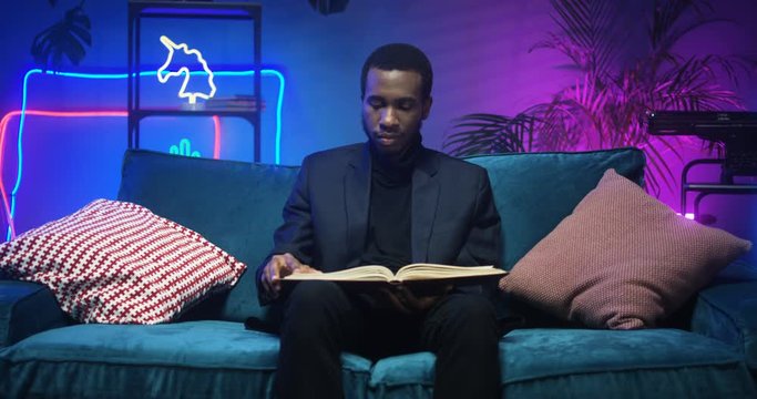 African American young stylish man sitting on sofa and reading book in living room with neon lights. Good looking black guy flipping pages of textbook on couch in futuristic atmosphere.
