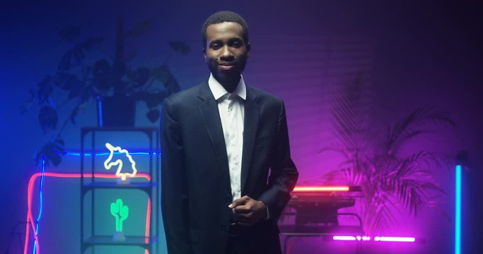 Young African American handsome gentleman in suit standing in room with blue and pink lights of neon tubes. Portrait of good looking man turning face to camera and smiling. Indoors.