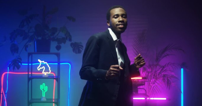 African American young handsome man in fancy suit dancing in room with neon lights. Black good looking gentleman at futuristic party. Guy in blue and pink lights of asteras. Male funny dancer. Indoor.