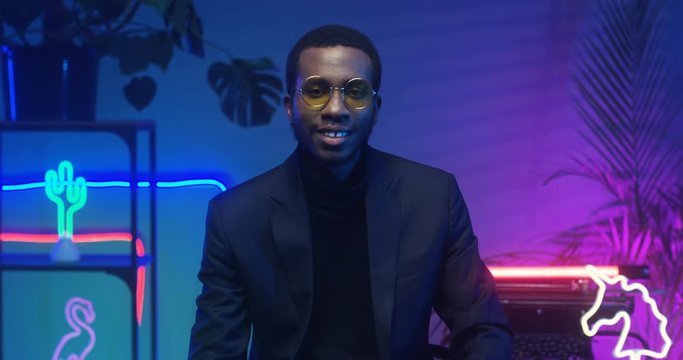 Portrait shot of African American handsome man in glasses looking straight at camera while sitting in futuristic room with blue and pink lights of neon tubes. Young guy smiling in club atmosphere.