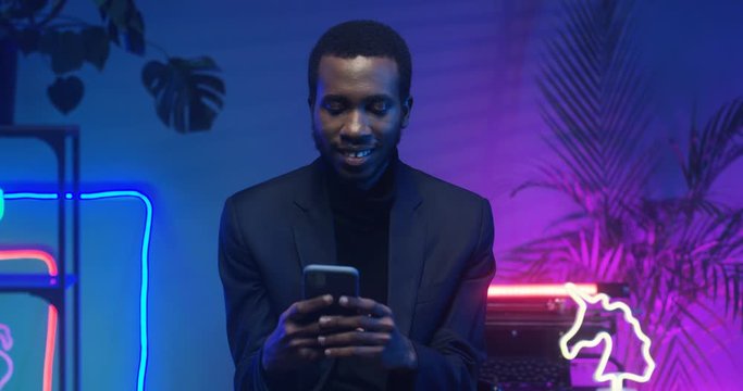 Smiled handsome and stylish African American man sitting. Black guy using smartphone in living room with neon lights. Male tapping, typing and scrolling on phone while chatting with messages.