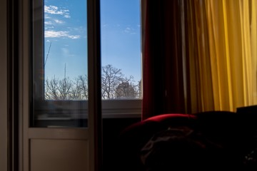 Afternoon sun on beautiful blue sky and clouds from inside the room with vibrant dark red and yellow drapes and dark red luxury material sofa behind open terrace door