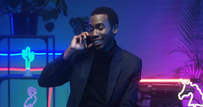 Smiled handsome and stylish African American man sitting and talking on cellphone. Black guy speaking on phone in living room with neon lights. Male having conversation on mobile telephone.