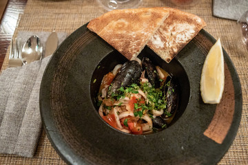 Roast mussels and squid with tomatoes and herbs in a dark plate with lemon and slices of bread. Delicacy