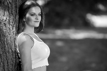 Black and white portrait of beautiful young woman in white tank top leaning against tree - subtle highlights and shadows
