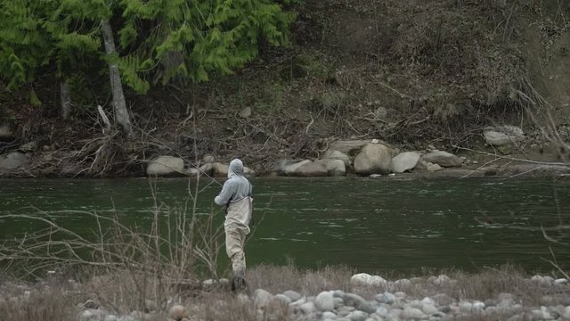 Male in waders alone fishing peacefully reeling in lure on river water bank on sandy river in pacific northwest oregon wide angle in slow motion