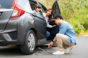 Fototapeta na wymiar Asian young father change changing the punctured tyre on his car loosening the nuts with a wheel spanner before jacking up the vehicle and mother and daughter waiting between trip and accident.