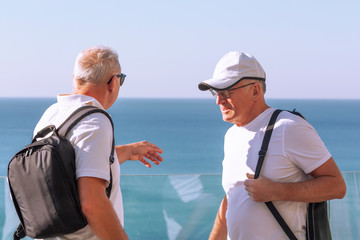 Two elderly tourists of athletic build talk on the background of the sea