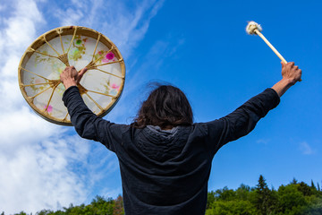 Rear view of long haired native american young man with sacred native frame drum and fur covered stick worshipping lake with arms stretched