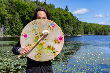Closeup of long haired native american young man holding sacred native frame drum with fur covered stick at lake with waterlilies covering face