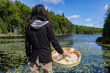 Rear view of long haired native young man standing holding sacred native frame drum near lake with...