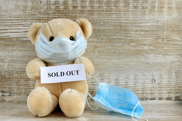 Announcement with text sold out held by a toy Teddy bear in a protective medical mask on a wooden...