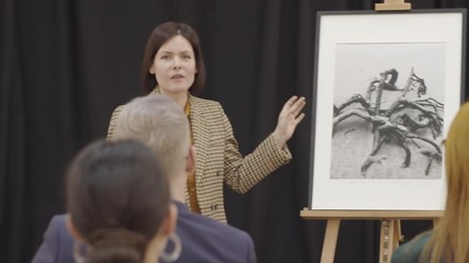 Attractive female auctioneer standing at lectern and presenting photography to audience at art auction