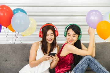 Two asian girls sitting together on sofa with headphone on and looking at a mobile phone device...