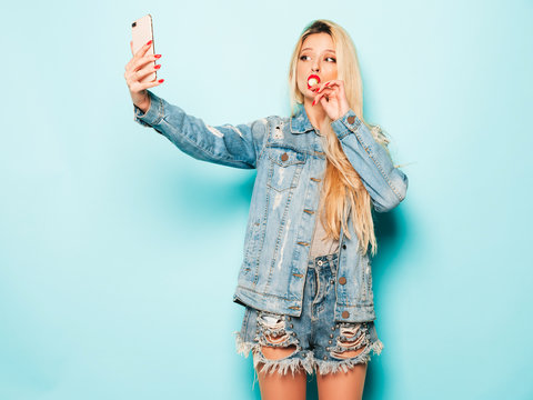 Young beautiful hipster bad girl in trendy jeans summer clothes and earring in her nose.Sexy carefree smiling blond woman posing in studio.Positive model licking round sugar candy.Takes selfie photo
