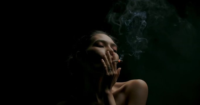Slow motion portrait of attractive young asian woman smoking a cigarette in low light on black background.