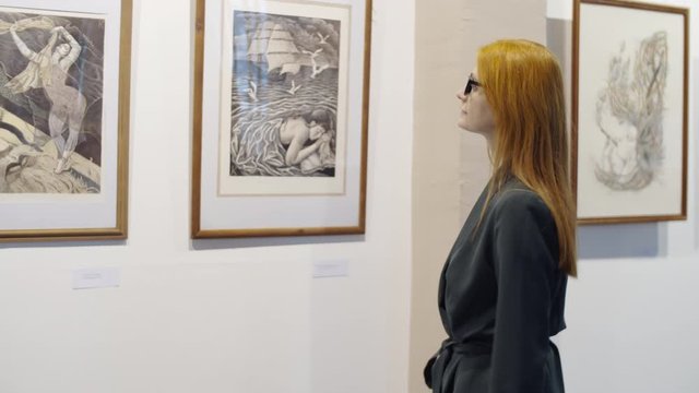 Elegant redhead woman in eyeglasses walking in art gallery and examining modern artworks on the wall