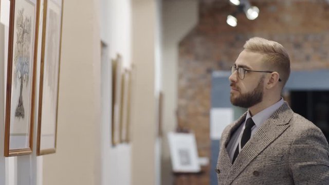Caucasian man in glasses and elegant outfit standing in art gallery, holding hand on chin, looking at artwork on the wall and trying to understand it