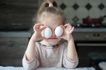 Little girl holds two eggs before her eyes in the kitchen