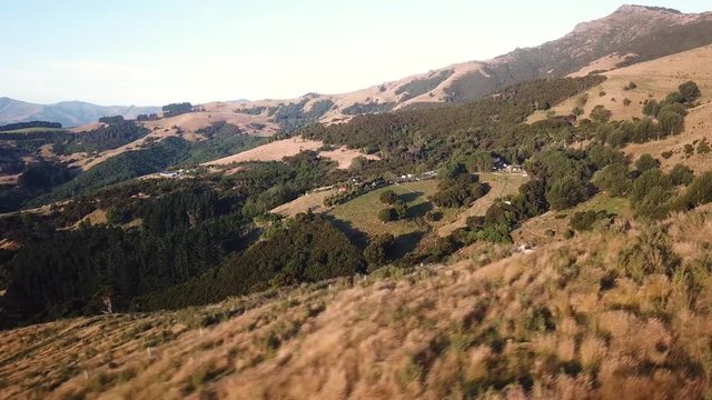 Aerial drone footage flying across the mountains of Akaroa over green hills and trees in new zealands south island during sunset.