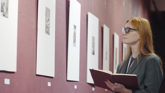Intelligent redhead woman in glasses looking at artwork in gallery and taking notes in notepad while writing review of exhibition