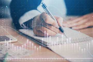 A woman hands writing information about stock market in notepad. Forex chart holograms in front....