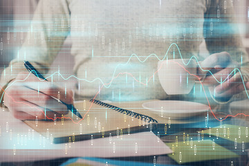 Forex chart hologram on hand taking notes background. Concept of analysis. Double exposure