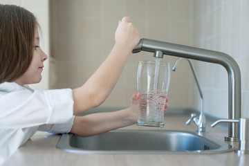 Portrait of a little caucasian girl gaining a glass of tap clean water. Kitchen faucet. Cute curly...