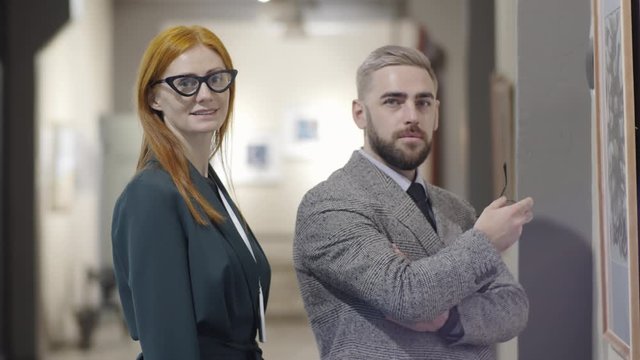 Waist up shot of beautiful redhead woman and handsome elegant man standing together in art gallery, posing for camera and smiling