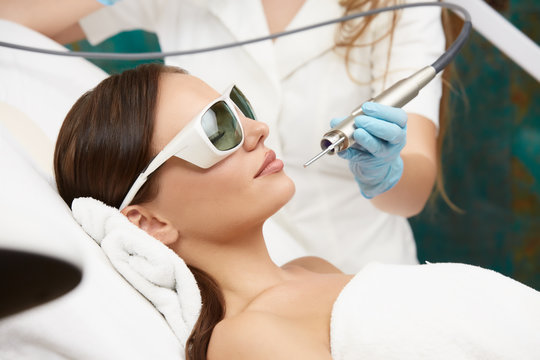 relaxed woman in cosmetology clinic wearing white bathrobe with beautician holding laser near her face