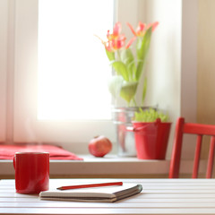 mug on the table with a pencil and a notebook on the background of a window with flowers in the morning. coffee-warming pause in red colors