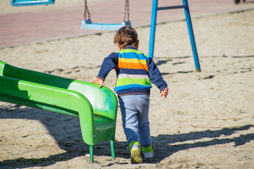 Little boy dressed in colorful clothes plays in the playground. The child plays on the slide. Rear view.