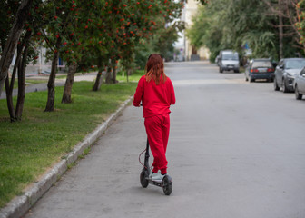 Red-haired girl in a red tracksuit drives an electric scooter. A young woman in oversized clothes rides around the city and listens to music using wireless headphones. View from the back.