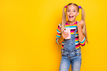 Surprised crazy energetic preteen kid use smartphone read social media news win lottery raise fists scream yes wear striped sweater jumper denim jeans isolated yellow bright color background