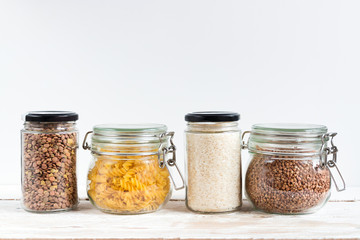 A variety of crops (buckwheat, rice, pasta, chickpeas) in glass jars on a wooden table on a light background. Close-up. The concept of vegetarianism and a balanced diet.