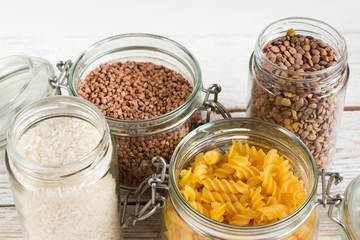 A variety of crops (buckwheat, rice, pasta, chickpeas) in glass jars on a wooden table on a light background. Close-up. The concept of vegetarianism and a balanced diet.