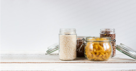 A variety of crops (buckwheat, rice, pasta, chickpeas) in glass jars on a wooden table on a light background. Close-up. The concept of vegetarianism and a balanced diet.